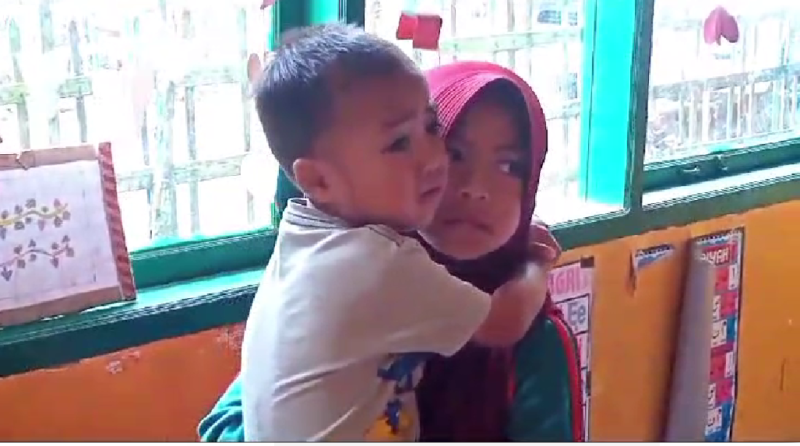 Foto : Nuraeni saat (9) menggendong adiknya saat belajar di sekolah